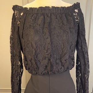 May include: Black jumpsuit with a lace off-the-shoulder top and wide-leg pants. The top features a delicate floral lace pattern, and the pants have a high waist. The jumpsuit is displayed on a mannequin.