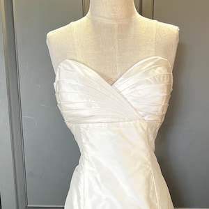 May include: A strapless white wedding dress on a mannequin. The bodice features a ruched design, and the skirt flows to the floor. The dress is made of a shiny, satin-like fabric. The background includes a gray cabinet and a colorful rug.