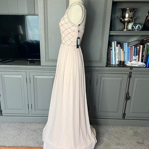 Puede incluir: Un vestido de noche rosa p&aacute;lido exhibido en un maniqu&iacute;. El corpi&ntilde;o presenta un dise&ntilde;o de cuentas y la falda llega hasta el suelo. Una etiqueta cuelga del vestido. El vestido se exhibe frente a un armario gris y una alfombra estampada.