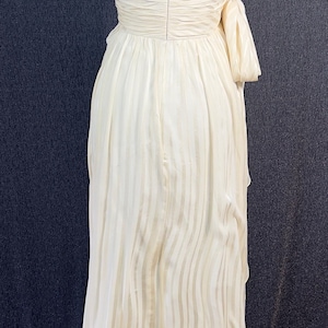 May include: A floor-length cream-colored strapless dress featuring a vertical striped pattern, a fitted bodice with a center back zipper, and a gathered waistline with a decorative side bow detail. The skirt flows downward in a straight silhouette.