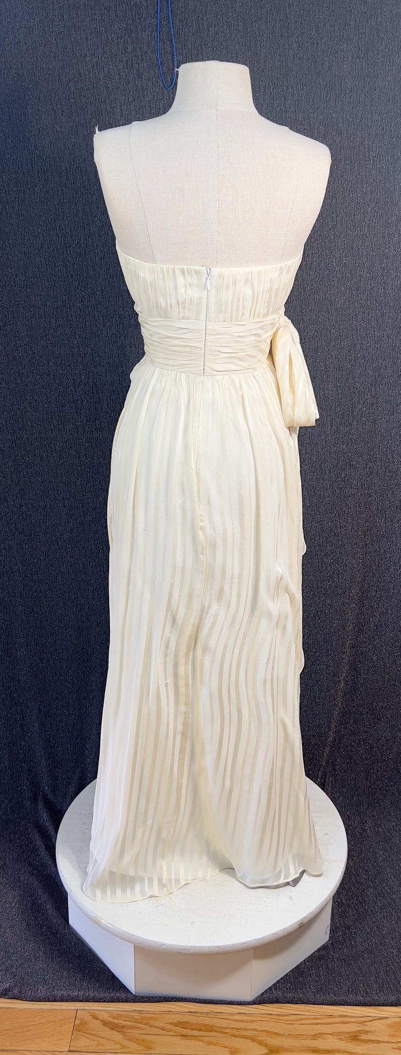 May include: A floor-length cream-colored strapless dress featuring a vertical striped pattern, a fitted bodice with a center back zipper, and a gathered waistline with a decorative side bow detail. The skirt flows downward in a straight silhouette.
