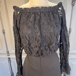 Puede incluir: Mono negro con hombros descubiertos. La parte superior est&aacute; hecha de intrincado encaje negro con mangas largas y un escote con volantes. La parte inferior es de color negro liso, de dise&ntilde;o de pierna ancha. El mono se muestra en un maniqu&iacute;.