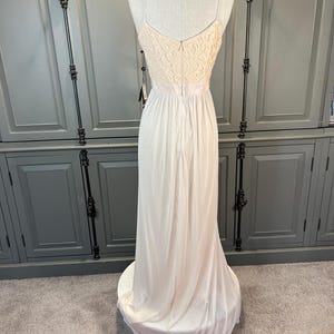 May include: A floor-length, ivory-colored gown displayed on a mannequin. The bodice features a cream-colored lace design, with spaghetti straps and a low back. The skirt flows to the floor, creating a soft, elegant silhouette. A tag hangs from the dress.