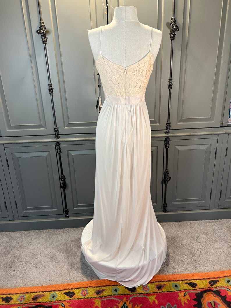 May include: A floor-length, ivory-colored gown displayed on a mannequin. The bodice features a cream-colored lace design, with spaghetti straps and a low back. The skirt flows to the floor, creating a soft, elegant silhouette. A tag hangs from the dress.