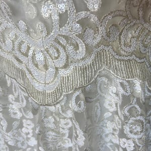 May include: White lace fabric with a delicate floral pattern and sequins. The fabric has a soft, flowing texture and is perfect for creating garments or accessories.