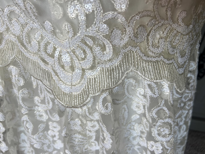 May include: White lace fabric with a delicate floral pattern and sequins. The fabric has a soft, flowing texture and is perfect for creating garments or accessories.