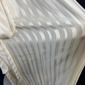 May include: A piece of off-white fabric featuring a vertical striped pattern. The material has a subtle sheen and a textured surface with alternating opaque and semi-sheer stripes running parallel along the length of the draped textile.