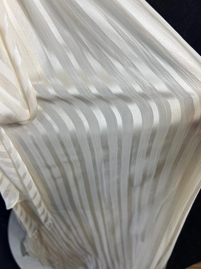 May include: A piece of off-white fabric featuring a vertical striped pattern. The material has a subtle sheen and a textured surface with alternating opaque and semi-sheer stripes running parallel along the length of the draped textile.