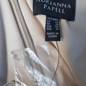 May include: Close-up of a dress tag from Adrianna Papell. The tag is black with white text, showing size information for US, EU, UK, GER, and AUS. The tag also states "MADE IN CHINA". A zipper and fabric are visible.