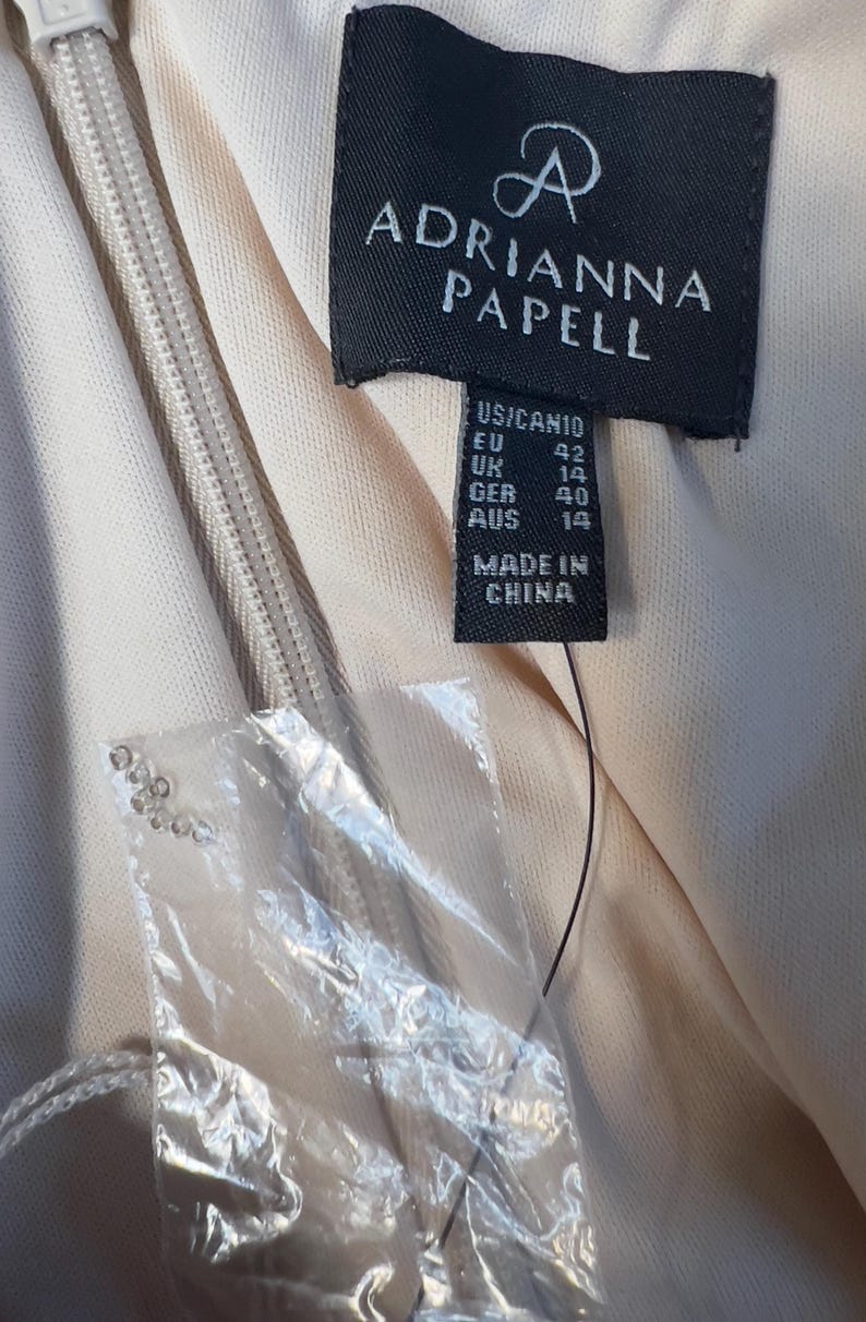 May include: Close-up of a dress tag from Adrianna Papell. The tag is black with white text, showing size information for US, EU, UK, GER, and AUS. The tag also states "MADE IN CHINA". A zipper and fabric are visible.