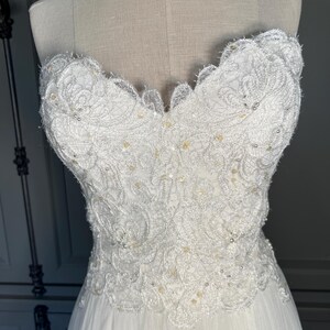 May include: A strapless white wedding dress with intricate lace detailing on the bodice. The bodice is embellished with small, scattered beads. The skirt is made of a flowing, sheer fabric.