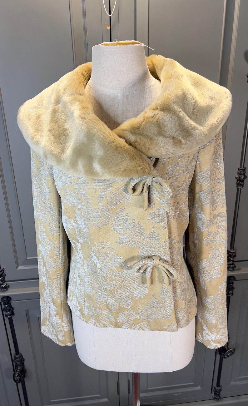 May include: A vintage-style jacket with a cream-colored floral pattern and a faux fur collar. The jacket features bow closures and long sleeves. The jacket is displayed on a mannequin.