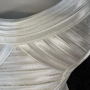May include: Close-up of a white, form-fitting dress. The dress features horizontal bands of fabric with a ribbed texture, creating a structured look. The fabric appears to be a satin or similar material, with a slight sheen. The dress has a scoop neckline.