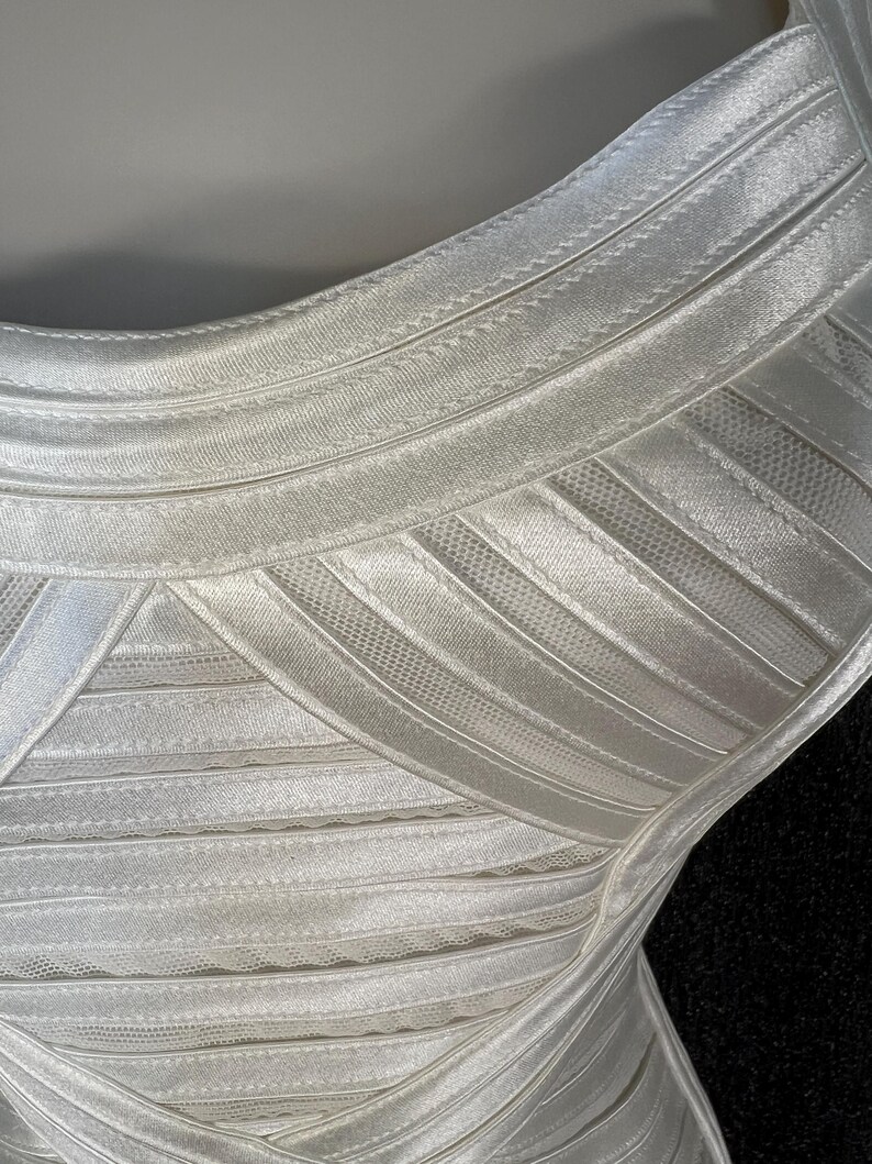 May include: Close-up of a white, form-fitting dress. The dress features horizontal bands of fabric with a ribbed texture, creating a structured look. The fabric appears to be a satin or similar material, with a slight sheen. The dress has a scoop neckline.