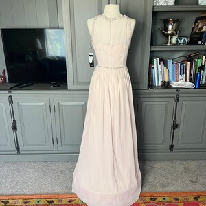 Puede incluir: Vestido de noche rosa p&aacute;lido sobre un maniqu&iacute;. El vestido presenta un escote con cuentas y una falda fluida. Un cintur&oacute;n fino con cuentas acent&uacute;a la cintura. El vestido es largo hasta el suelo y parece estar hecho de una tela ligera.
