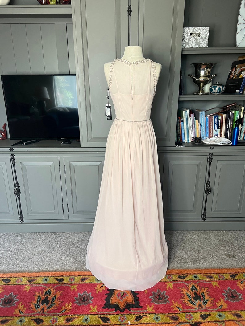 Puede incluir: Vestido de noche rosa p&aacute;lido sobre un maniqu&iacute;. El vestido presenta un escote con cuentas y una falda fluida. Un cintur&oacute;n fino con cuentas acent&uacute;a la cintura. El vestido es largo hasta el suelo y parece estar hecho de una tela ligera.