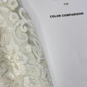 May include: A close-up of a white fabric with a floral pattern made of sequins. The fabric is draped over a white piece of paper with the text "STANDARD WHITE PRINTER PAPER FOR COLOR COMPARISON".