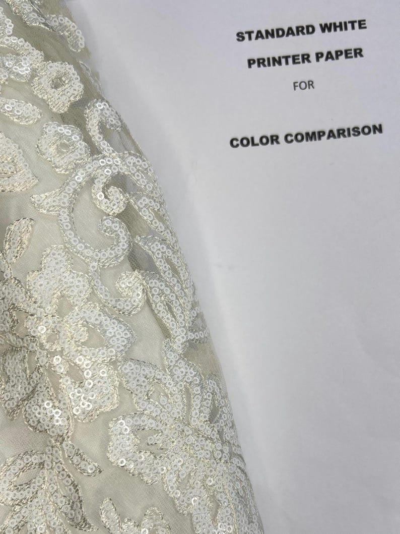 May include: A close-up of a white fabric with a floral pattern made of sequins. The fabric is draped over a white piece of paper with the text "STANDARD WHITE PRINTER PAPER FOR COLOR COMPARISON".