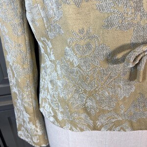 May include: Close-up of a tan jacket with a floral pattern in shades of white and gray. The jacket has long sleeves and a tie detail at the waist. The fabric has a textured appearance.