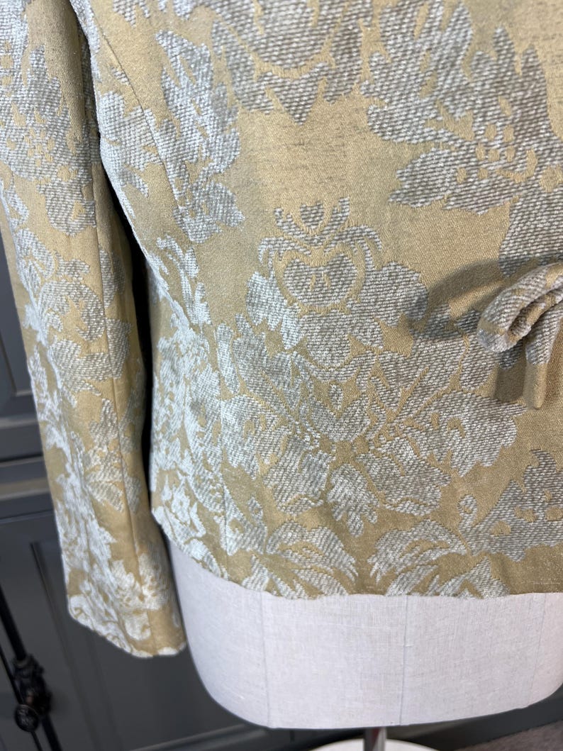 May include: Close-up of a tan jacket with a floral pattern in shades of white and gray. The jacket has long sleeves and a tie detail at the waist. The fabric has a textured appearance.