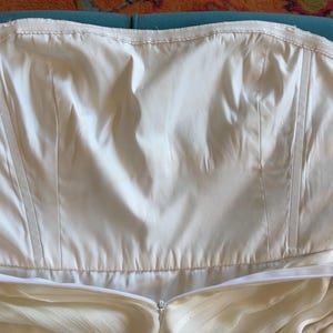 May include: A strapless white bodice featuring vertical boning channels and a smooth, opaque fabric construction. The garment includes a horizontal zipper closure along the lower edge, with a clean, structured design and a plain, unadorned surface.