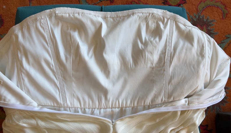 May include: A strapless white bodice featuring vertical boning channels and a smooth, opaque fabric construction. The garment includes a horizontal zipper closure along the lower edge, with a clean, structured design and a plain, unadorned surface.