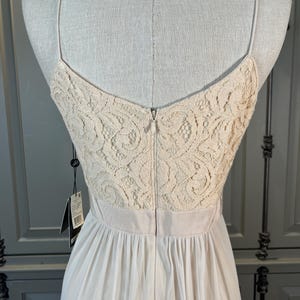 May include: Back view of a cream-colored dress on a mannequin. The bodice features a delicate lace design, transitioning to a flowing, pleated skirt. The dress has thin spaghetti straps and a zipper closure down the back. A tag hangs from the dress.