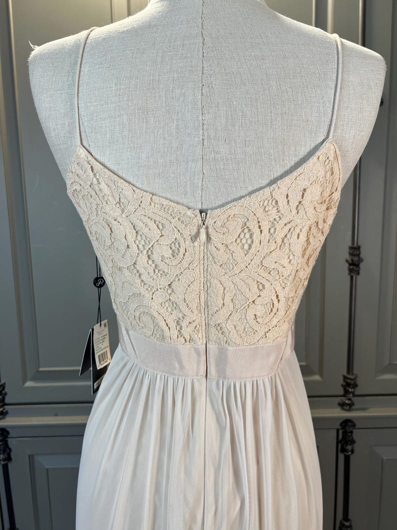 May include: Back view of a cream-colored dress on a mannequin. The bodice features a delicate lace design, transitioning to a flowing, pleated skirt. The dress has thin spaghetti straps and a zipper closure down the back. A tag hangs from the dress.