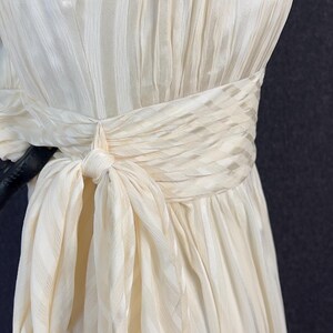May include: A strapless cream-colored dress featuring a subtle vertical striped texture. The garment includes a wide, gathered waistband that wraps around the midsection and ties into a prominent bow on the side, creating a draped, pleated effect.