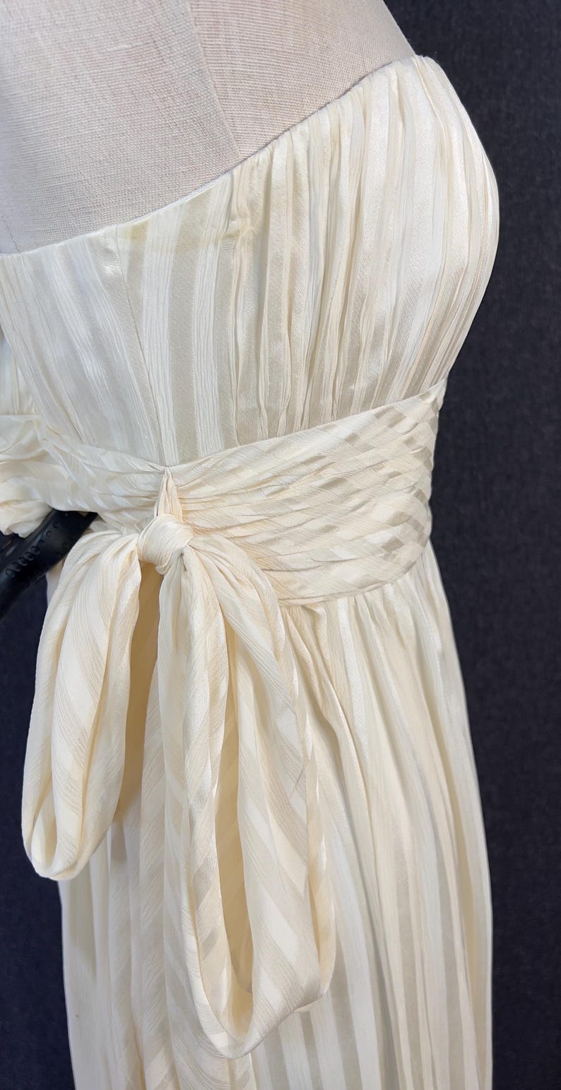 May include: A strapless cream-colored dress featuring a subtle vertical striped texture. The garment includes a wide, gathered waistband that wraps around the midsection and ties into a prominent bow on the side, creating a draped, pleated effect.