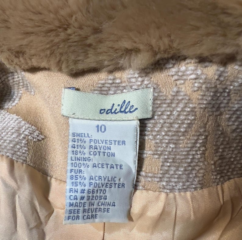 May include: Close-up of a tan and cream-colored jacket with a faux fur collar. The label reads "odille" and lists the fabric composition: 41% polyester, 41% rayon, and 18% cotton. The lining is 100% acetate, and the fur is 85% acrylic and 15% polyester.