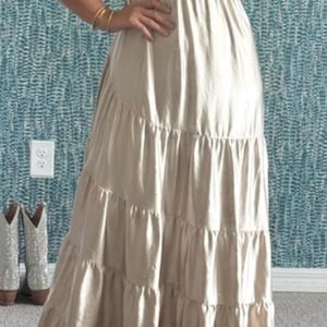 May include: A beige maxi dress with a tiered skirt and puffed sleeves. The dress has a sweetheart neckline and a fitted bodice. The model is wearing black heels and gold bracelets. A pair of cowboy boots are in the background.
