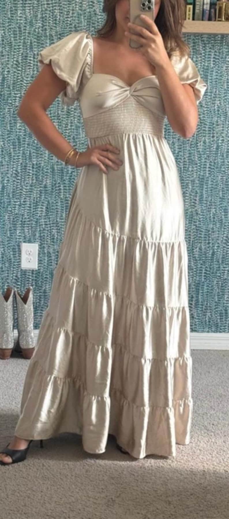 May include: A beige maxi dress with a tiered skirt and puffed sleeves. The dress has a sweetheart neckline and a fitted bodice. The model is wearing black heels and gold bracelets. A pair of cowboy boots are in the background.