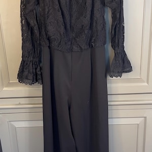 Puede incluir: Mono negro con top de encaje con hombros descubiertos y pantal&oacute;n de pernera ancha. La parte superior tiene mangas largas con pu&ntilde;os de encaje acampanados. La prenda cuelga de una percha blanca sobre un fondo neutro.