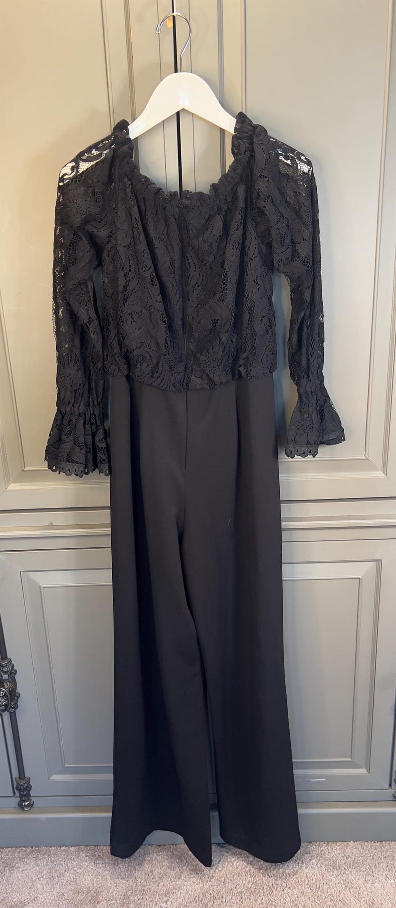 Puede incluir: Mono negro con top de encaje con hombros descubiertos y pantal&oacute;n de pernera ancha. La parte superior tiene mangas largas con pu&ntilde;os de encaje acampanados. La prenda cuelga de una percha blanca sobre un fondo neutro.