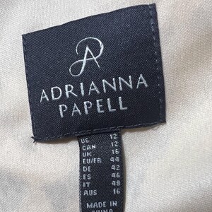 Op de afbeelding: Zwart-witte maatlabel met de tekst "ADRIANNA PAPELL" en maatconversies voor de VS, Canada, VK, EU/FR, DE, ES, IT en AUS. Het label vermeldt ook "MADE IN CHINA".