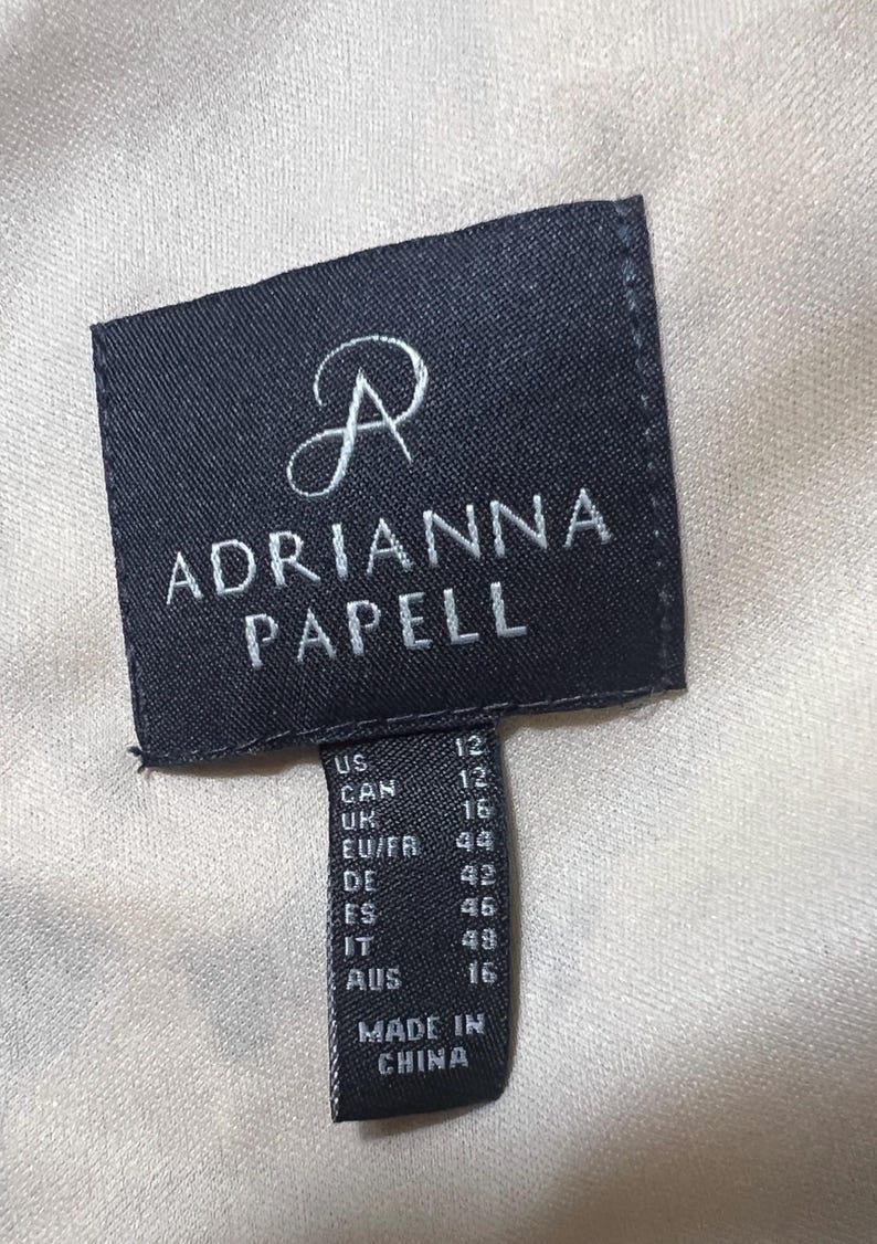 Op de afbeelding: Zwart-witte maatlabel met de tekst "ADRIANNA PAPELL" en maatconversies voor de VS, Canada, VK, EU/FR, DE, ES, IT en AUS. Het label vermeldt ook "MADE IN CHINA".