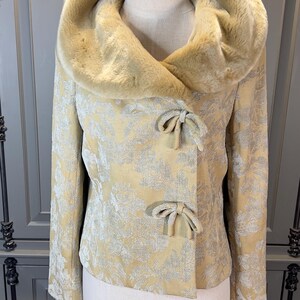 May include: A vintage-style jacket with a faux fur collar and a floral pattern. The jacket is a light yellow color with silver accents and bow closures. The collar is a lighter shade of yellow.