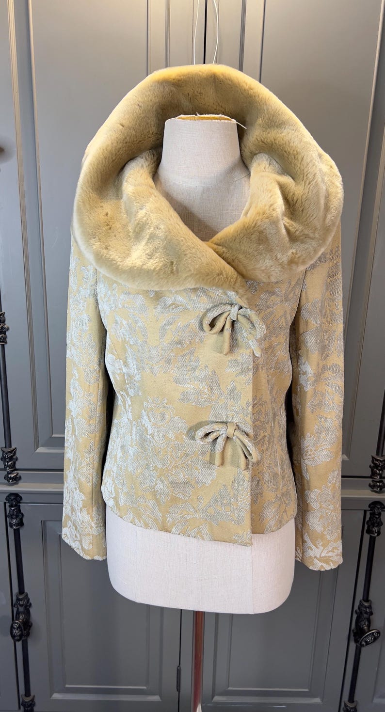 May include: A vintage-style jacket with a faux fur collar and a floral pattern. The jacket is a light yellow color with silver accents and bow closures. The collar is a lighter shade of yellow.