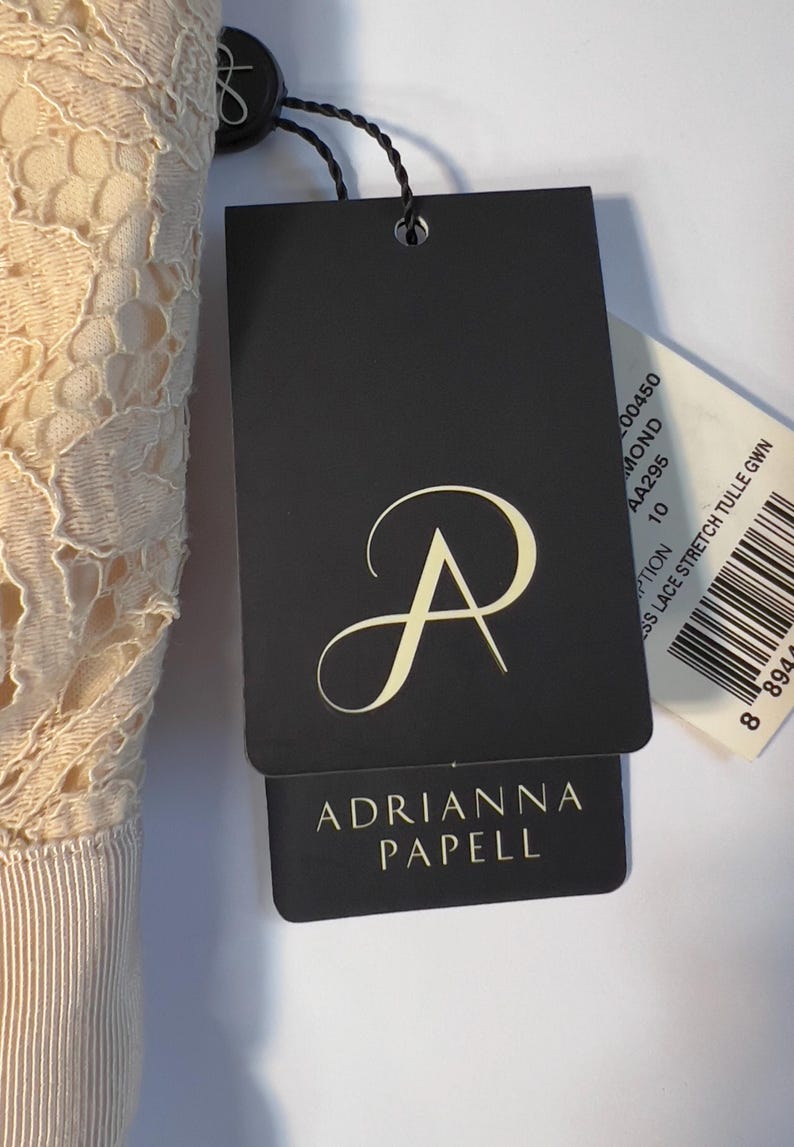 May include: A close-up of a cream-colored lace dress with a tag from Adrianna Papell. The tag is black with the brand's logo and name in gold. A smaller tag with a barcode is also visible.