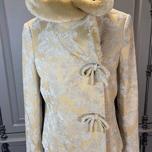 May include: A gold-colored jacket with a floral pattern and a faux fur collar. The jacket features bow closures and a tailored fit. The jacket is displayed on a mannequin.