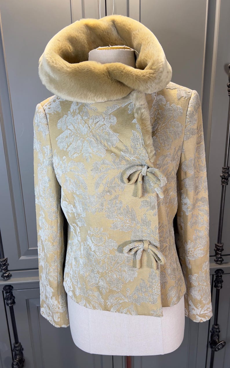 May include: A gold-colored jacket with a floral pattern and a faux fur collar. The jacket features bow closures and a tailored fit. The jacket is displayed on a mannequin.