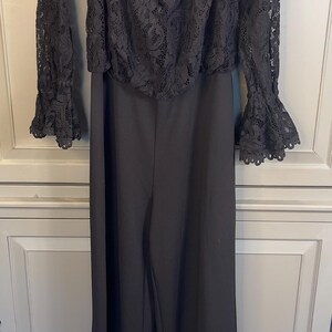Puede incluir: Mono negro con top de encaje con hombros descubiertos y pantal&oacute;n de pernera ancha. La parte superior tiene mangas largas con pu&ntilde;os acampanados. La prenda est&aacute; colgada de una percha blanca sobre un fondo neutro.