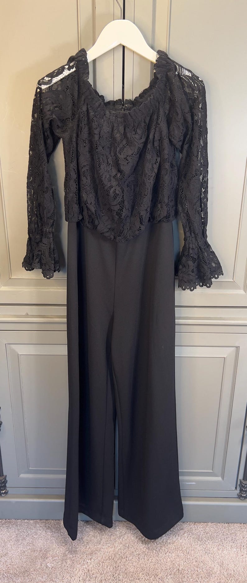 Puede incluir: Mono negro con top de encaje con hombros descubiertos y pantal&oacute;n de pernera ancha. La parte superior tiene mangas largas con pu&ntilde;os acampanados. La prenda est&aacute; colgada de una percha blanca sobre un fondo neutro.