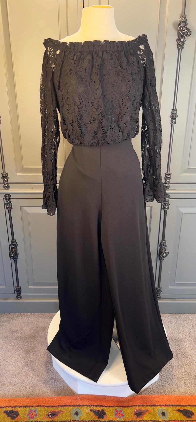 May include: Black jumpsuit with a lace off-the-shoulder top and wide-leg pants. The top features a delicate floral lace pattern, and the pants have a high waist. The jumpsuit is displayed on a mannequin.