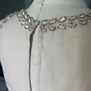 Puede incluir: Una blusa sin mangas de color rosa claro con un escote adornado con cuentas y lentejuelas. Los adornos plateados y transparentes a&ntilde;aden un toque de brillo. La tela parece ser gasa o seda ligera, con una textura sutil. La blusa se muestra en un maniqu&iacute;.