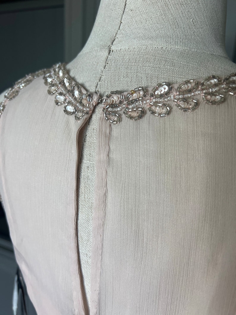 Puede incluir: Una blusa sin mangas de color rosa claro con un escote adornado con cuentas y lentejuelas. Los adornos plateados y transparentes a&ntilde;aden un toque de brillo. La tela parece ser gasa o seda ligera, con una textura sutil. La blusa se muestra en un maniqu&iacute;.