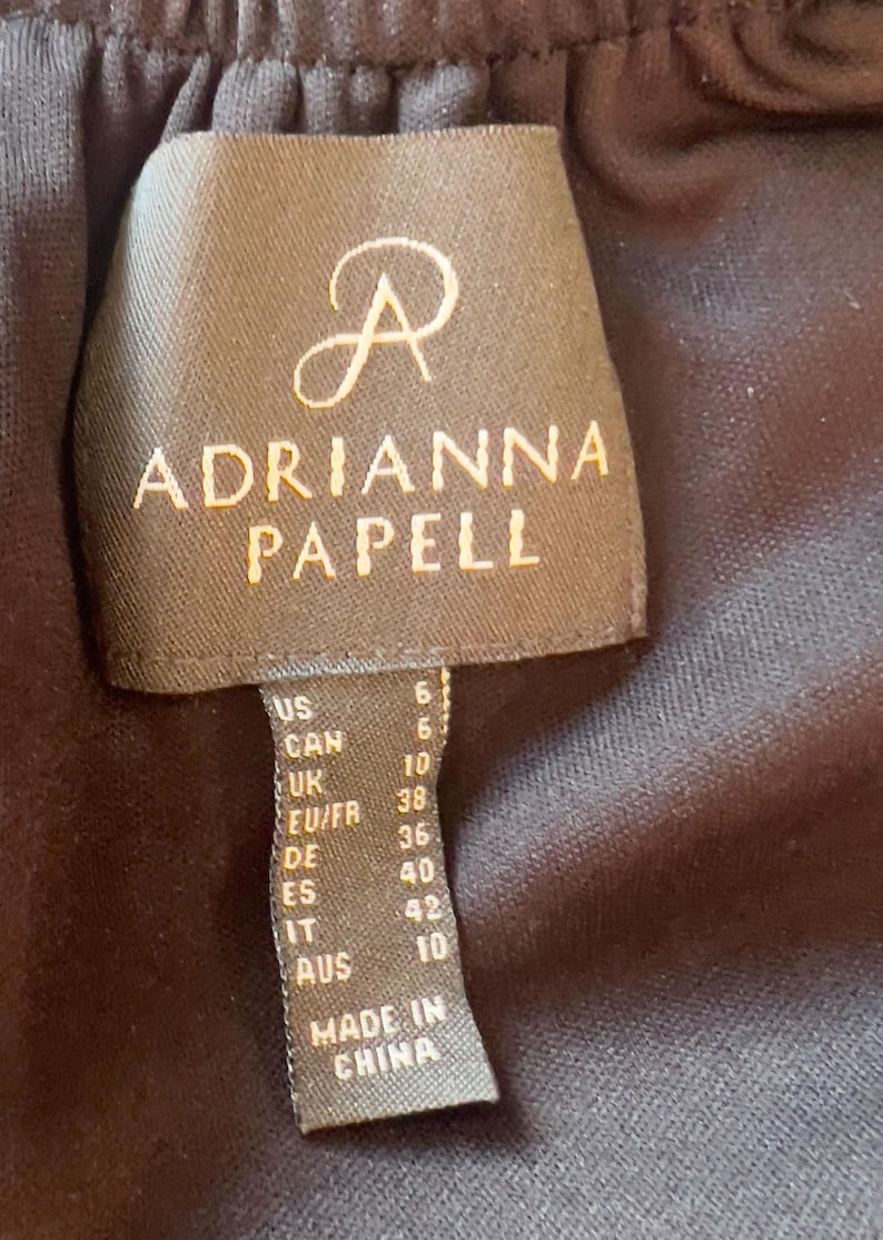 Puede incluir: Etiqueta negra de Adrianna Papell con informaci&oacute;n de tallas. La etiqueta muestra la talla US 6, CAN 6, UK 10, EU/FR 38, DE 36, ES 40, IT 42 y AUS 10. La etiqueta est&aacute; hecha en China.