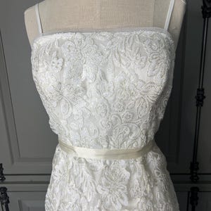 May include: A white sleeveless dress with a delicate floral pattern and a satin sash. The dress is made of a sheer fabric with sequins.