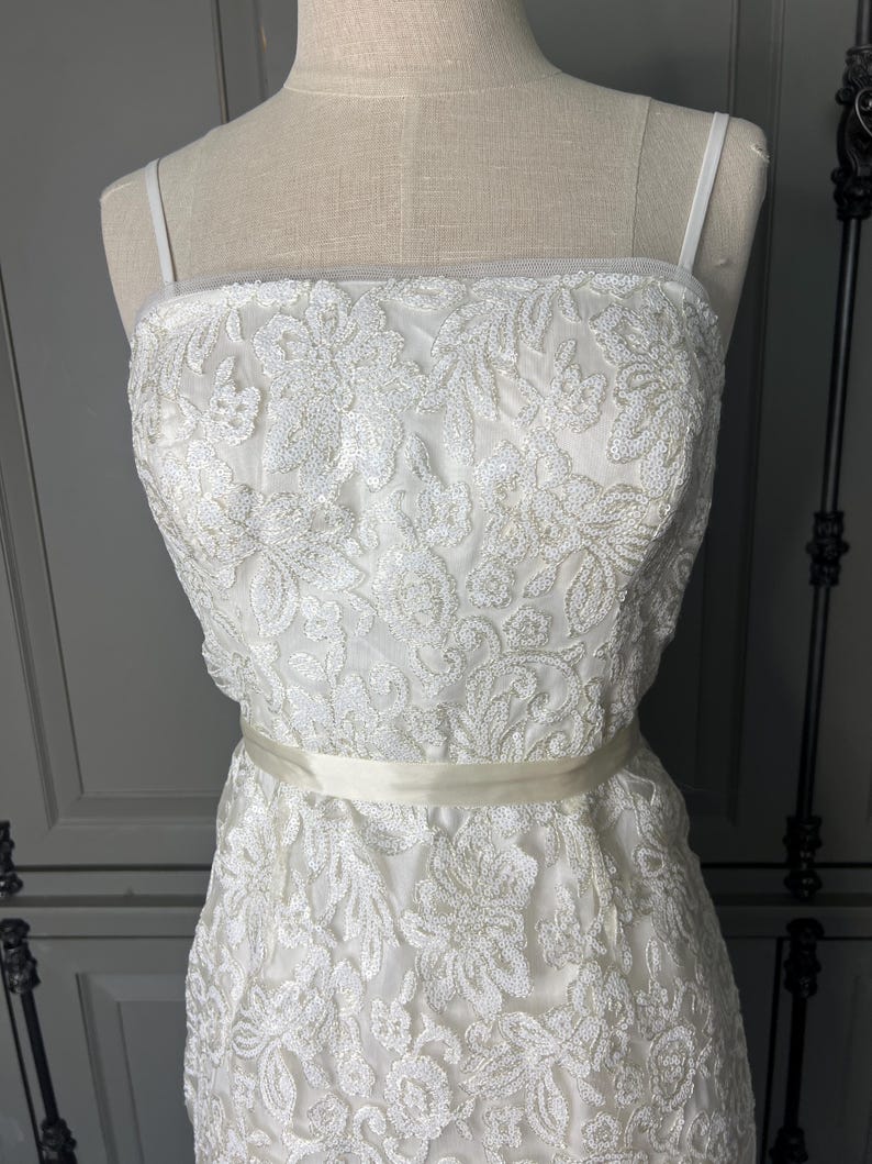 May include: A white sleeveless dress with a delicate floral pattern and a satin sash. The dress is made of a sheer fabric with sequins.